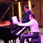 Pianist Ian Gindes sits at the piano with composer Patrick Zimmerli standing nearby as they together examine the musical score during rehearsals at Pianoforte Studios, Chicago.