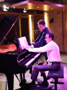 Pianist Ian Gindes sits at the piano with composer Patrick Zimmerli standing nearby as they together examine the musical score during rehearsals at Pianoforte Studios, Chicago.