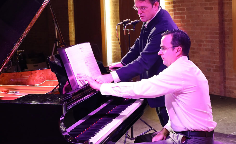 Pianist Ian Gindes sits at the piano with composer Patrick Zimmerli standing nearby as they together examine the musical score during rehearsals at Pianoforte Studios, Chicago.